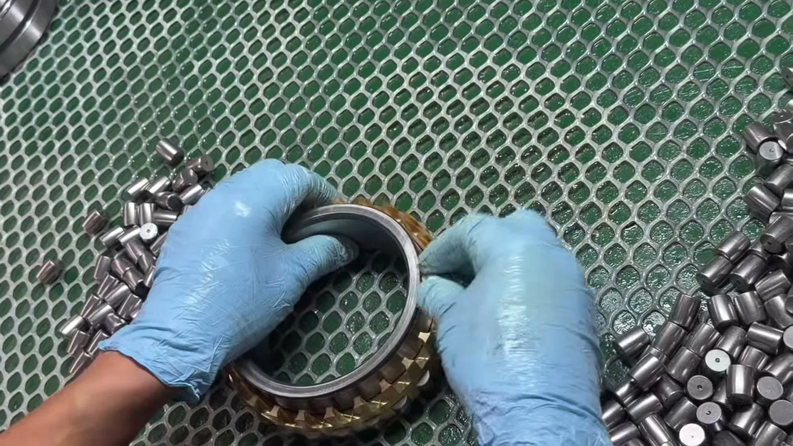 Gloved hands holding a roller bearing ring with a brass cage over a green mat, loose rollers beside it