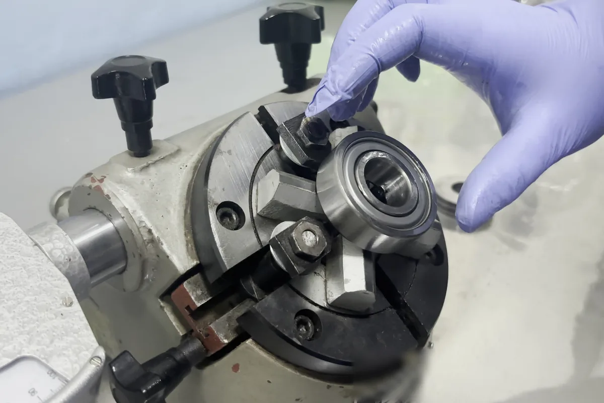 A gloved hand setting a small ball bearing into a chuck fixture on a bench