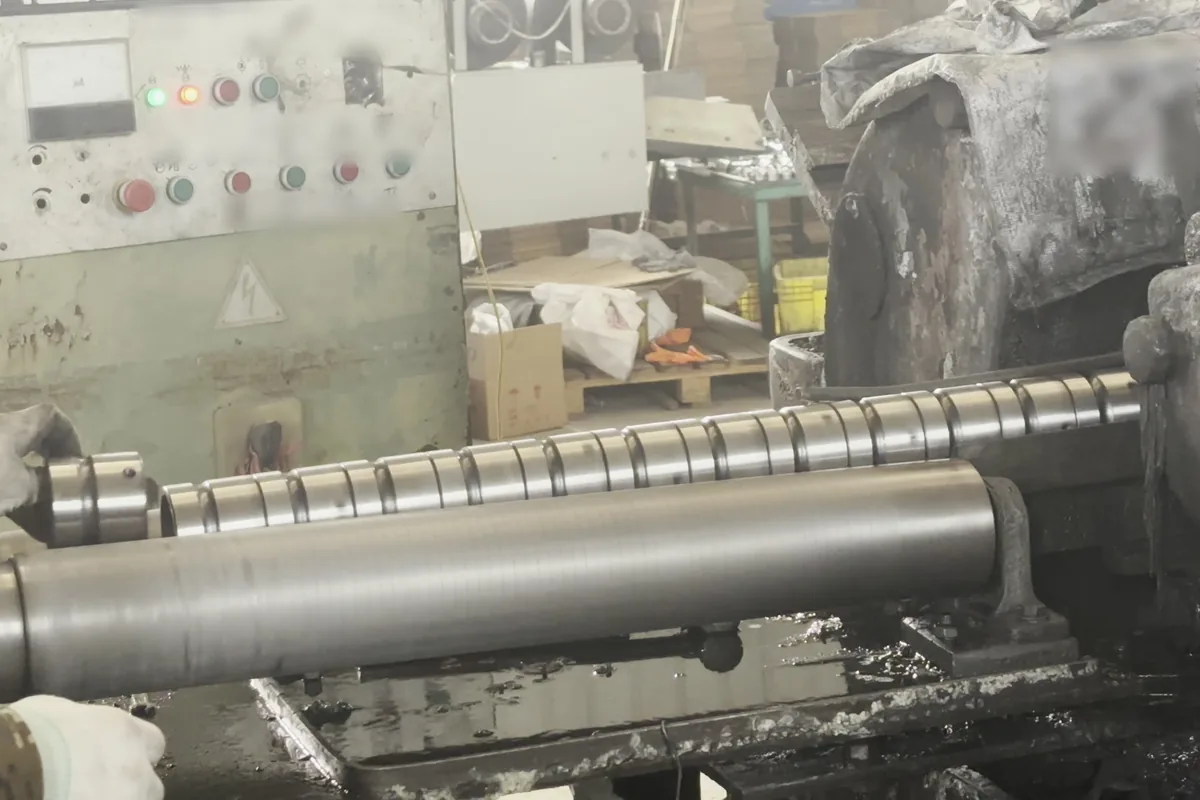 A long steel tube with cut grooves turning in a lathe, gloved hands at each end